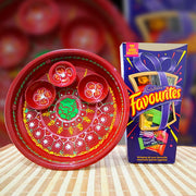 A red decorative clay diya set with painted floral patterns and a green center, shown next to its retail box labeled 'Candy Favourites' on a striped surface with a blurred red background.