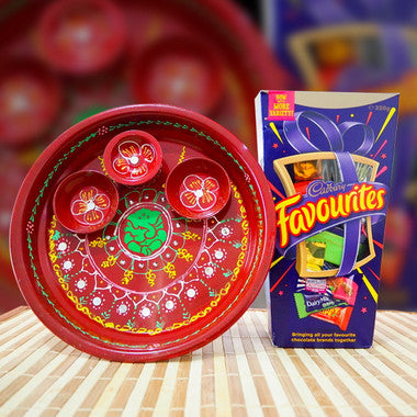 A red decorative clay diya set with painted floral patterns and a green center, shown next to its retail box labeled 'Candy Favourites' on a striped surface with a blurred red background.